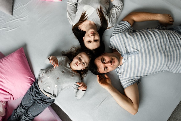 Bovenaanzicht van een schattig klein meisje en haar mooie jonge ouders die naar de camera kijken en glimlachen, thuis op de vloer liggen, Vvew van bovenaf van rustende familie die op de vloer ligt en naar de camera kijkt