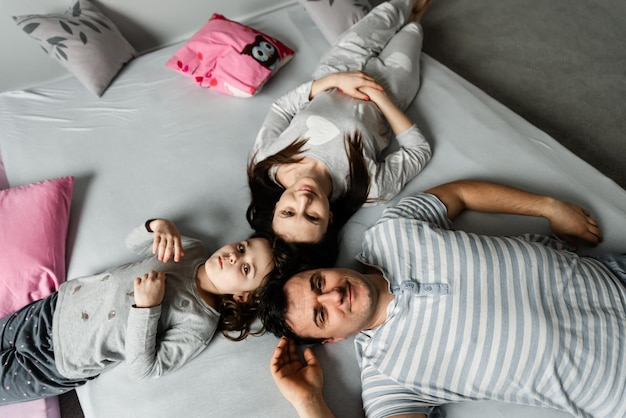 Bovenaanzicht van een schattig klein meisje en haar mooie jonge ouders die naar de camera kijken en glimlachen, thuis op de vloer liggen, Vvew van bovenaf van rustende familie die op de vloer ligt en naar de camera kijkt