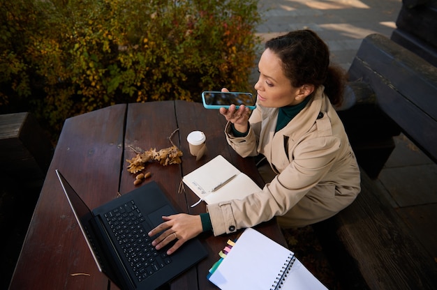 Bovenaanzicht van een Spaanse mooie vrouw, zelfverzekerde zakenvrouw die in een houten buitencafetaria voor haar laptop zit, een smartphone vasthoudt en een spraakbericht opneemt