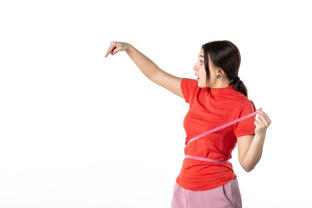 Bovenaanzicht van een verwarde jonge dame die haar haar verzamelt, gekleed in een oranje blouse en een meter vasthoudt die haar taille meet en naar beneden wijst op een witte achtergrond