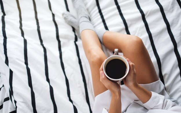 Bovenaanzicht van een vrouw die 's ochtends thuis warme koffie vasthoudt en drinkt in een wit, knus bed