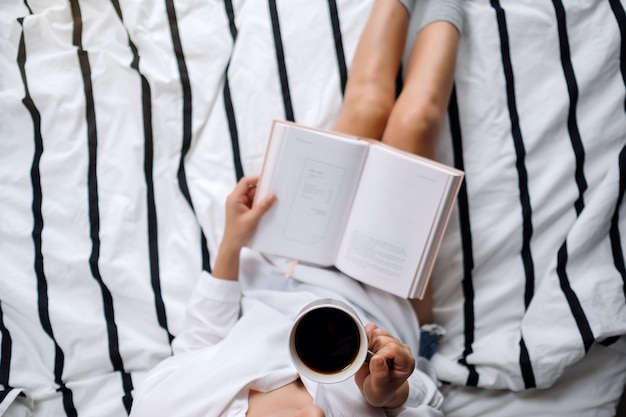 Bovenaanzicht van een vrouw leesboek en het drinken van warme koffie in een witte gezellige bed thuis in de ochtend