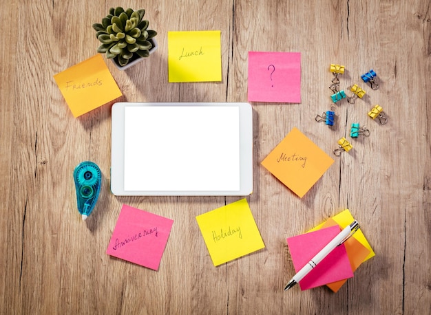 Bovenaanzicht van een witte digitale tablet met leeg scherm en veel zelfklevende notities met brainstorm op houten tafel.