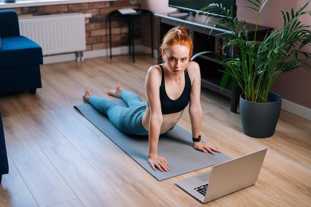 Bovenaanzicht van fit lachende roodharige jonge vrouw liggend op de vloer en het gebruik van laptopcomputer in appartement