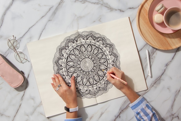 Foto bovenaanzicht van gekleurd mandala-kunstboek met kleurrijke markeringen, pen, roze etui en een kopje koffie