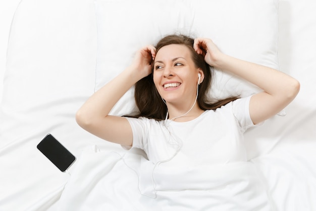 Bovenaanzicht van gelukkige jonge vrouw liggend in bed met wit laken, kussen, deken, luister muziek van oortelefoons in mobiele telefoon