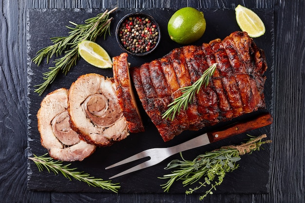 Bovenaanzicht van gesneden geroosterde varkensrollade - Porchetta, heerlijk gebraden varkensvlees van Italiaanse culinaire vakantietraditie op een lei dienblad met rozemarijn en limoen, close-up, flatlay