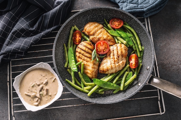 Bovenaanzicht van gezonde gekookte kip en groenten in een koekenpan met een romige champignonsaus aan de zijkant, geplaatst op een zwarte achtergrond met een handdoek en een pannenlap.