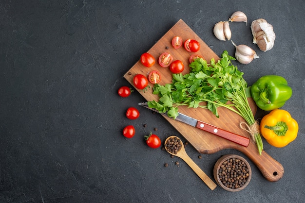 Bovenaanzicht van groene bundel verse hele gesneden tomaten op houten snijplank mes paprika's aan de linkerkant op zwarte noodlijdende oppervlak