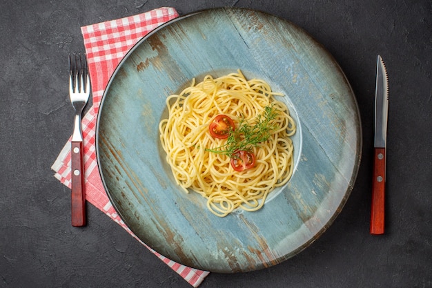 Bovenaanzicht van heerlijke spagetti geserveerd met tomatengroen en bestek op een rode gestripte handdoek op zwarte achtergrond