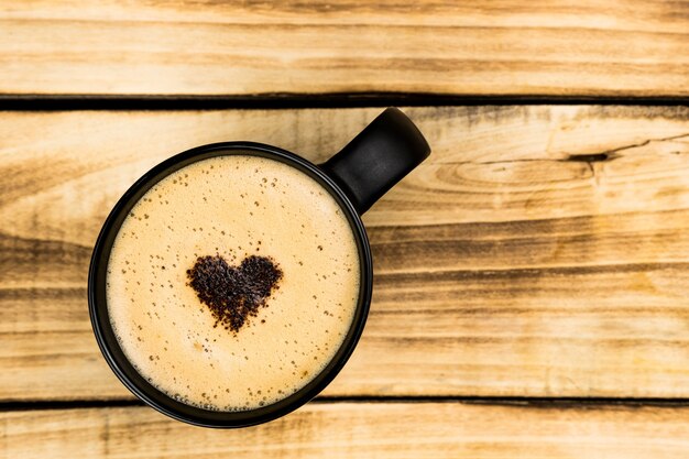 Bovenaanzicht van hete latte of cappuccino koffie in zwarte kop met latte art met hart binnen op houten tafel