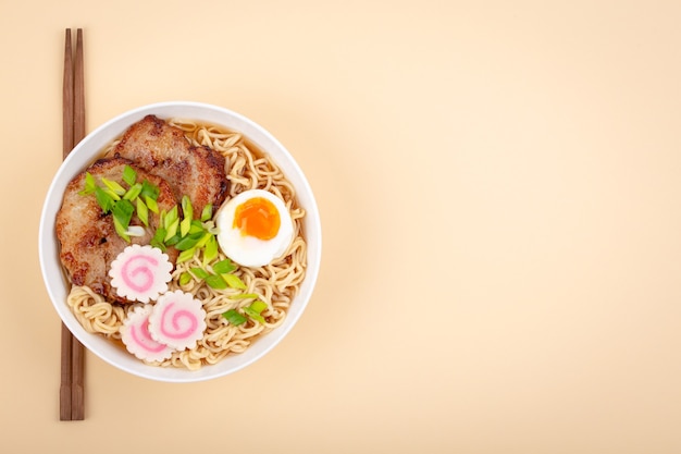 Bovenaanzicht van Japanse noedelsoep ramen in witte kom met noedels, vleesbouillon, gesneden geroosterd varkensvlees, narutomaki, ei met dooier op pastelbeige achtergrond. Traditioneel gerecht uit Japan, ruimte voor tekst