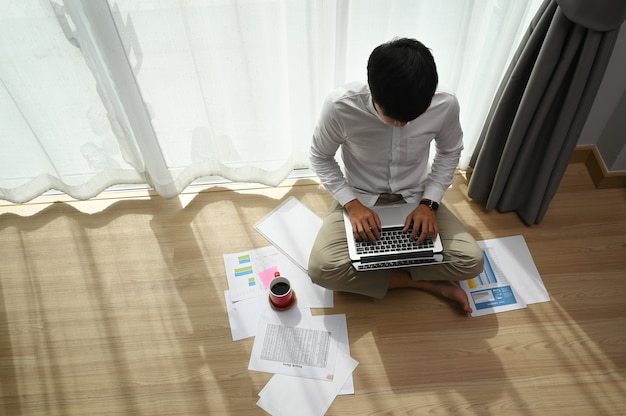 Bovenaanzicht van jonge man aan het werk te typen op laptop zittend op de vloer bij modern huis.