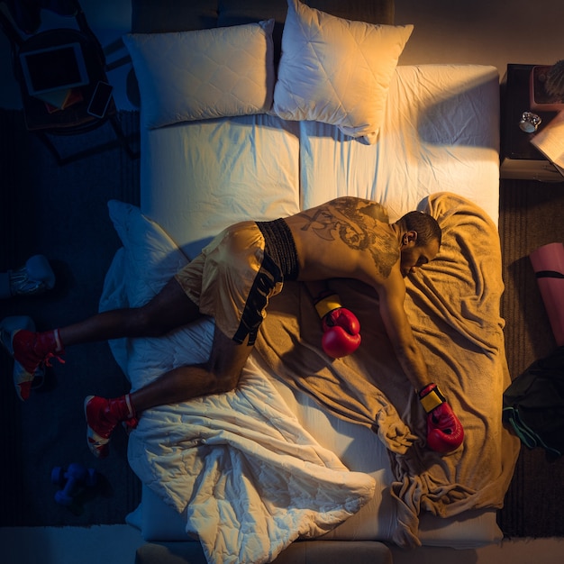 Bovenaanzicht van jonge professionele bokser, vechter die in zijn slaapkamer slaapt in sportkleding met handschoenen