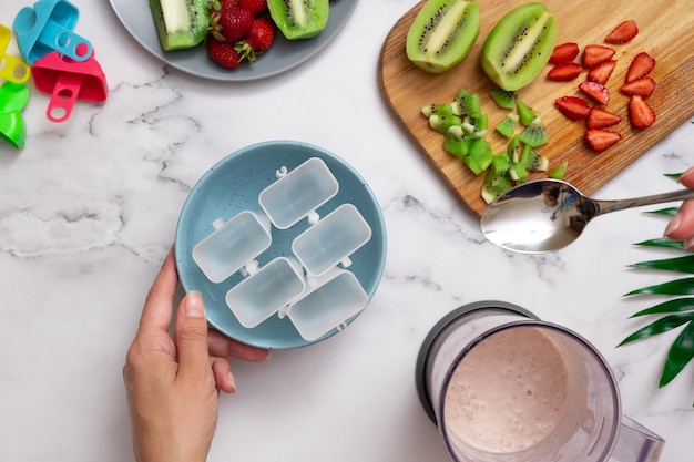 Bovenaanzicht van keukentafel met vrouwelijke handen die ijs in mallen gieten Zelfgemaakt ijs maken van yoghurt, aardbeien en kiwi