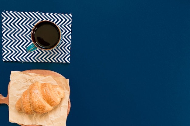 Bovenaanzicht van kopje zwarte koffie en croissant op een snijplank en ambachtelijke papier op blauwe achtergrond met kopie ruimte. Ochtendontbijt in Franse stijl.