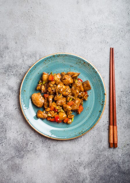 Bovenaanzicht van kung pao-kip op een bord klaar om te eten. gewokt chinees traditioneel gerecht met kip, pinda's, groenten, chilipepers. chinees diner, eetstokjes, rustieke betonnen achtergrond
