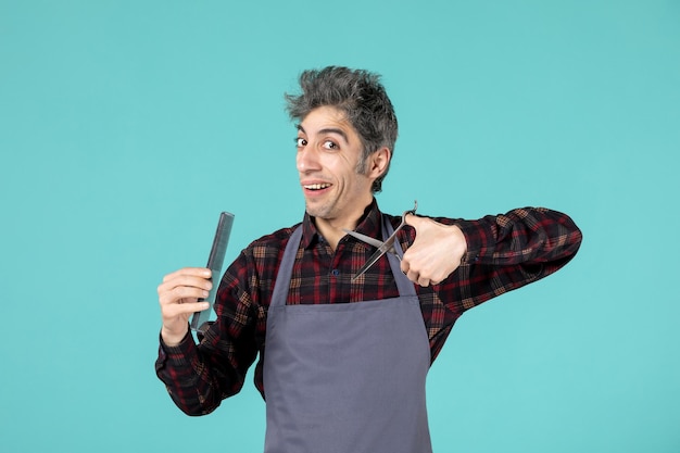 Bovenaanzicht van lachende gelukkige jonge kapper die een grijze schort draagt en een kam vasthoudt en een schaar toont op een pastelblauwe achtergrond