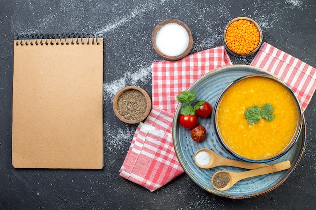 Foto bovenaanzicht van linzensoep in een kom geserveerd met groene tomaten peper zout op blauwe dienblad op rode gestripte handdoek en verschillende kruiden spiraal notebook op zwart witte achtergrond