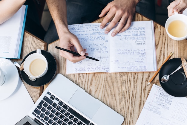 Bovenaanzicht van mensen die met copybook, boek en laptop studeren. Kopjes koffie op de tafel