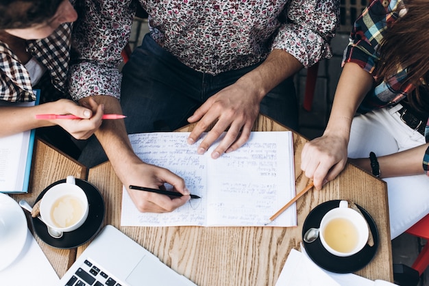 Bovenaanzicht van mensen die met copybook, boek en laptop studeren. Kopjes koffie op de tafel