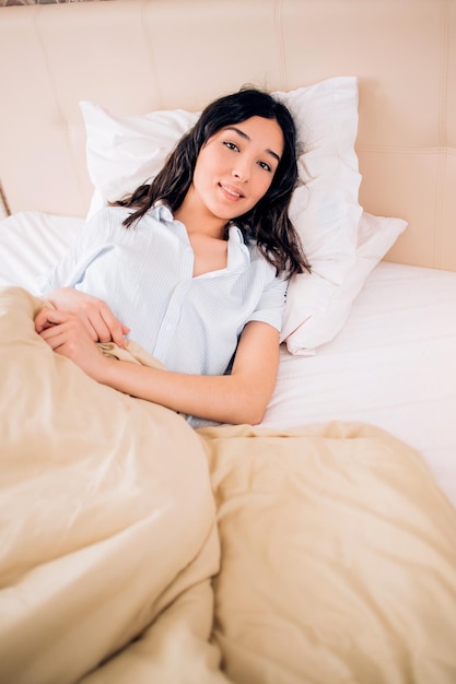 Bovenaanzicht van mooie jonge brunette vrouw liggend op bed blauw gestreepte pyjama dragen in modern appartement na het wakker worden. Ze bedekte met een warme deken, glimlachte naar de camera en zag er tevreden uit.
