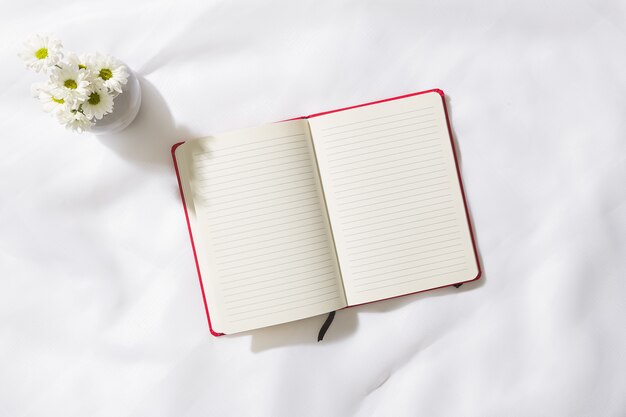 Bovenaanzicht van ochtendscène in voile stoffen achtergrond met een rood notitieboekje in het midden en een vaas met witte bloemen, met ruimte voor tekst