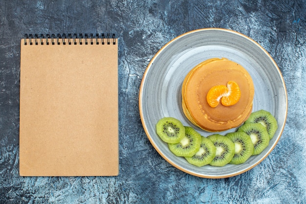 Bovenaanzicht van pluizige pannenkoeken in Amerikaanse stijl gemaakt met natuurlijke yoghurt geserveerd met kiwi's en mandarijn op een bord en notitieboekje op ijsachtergrond met vrije ruimte