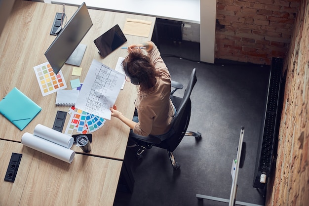 bovenaanzicht van professionele vrouwelijke ontwerper die een koptelefoon draagt met een blauwdruk terwijl hij aan het werk is