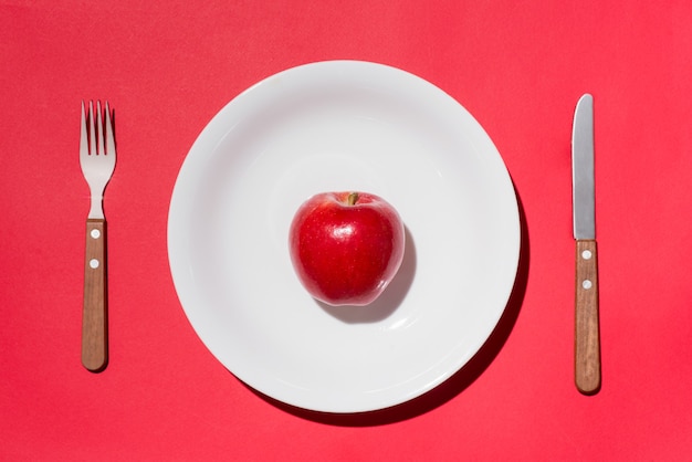 Bovenaanzicht van rode appel op witte plaat met mes en vork op rode achtergrond