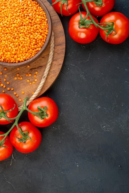 Bovenaanzicht van rode linzen in een bruine kom touw knoflook op houten ronde plank en tomaten met stengels op zwarte achtergrond