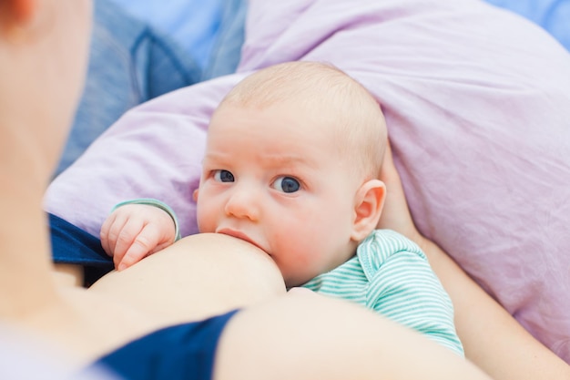 Bovenaanzicht van schattige baby borstvoeding, liggend op kussen onder moeders arm in rugbybalpositie, zachtjes ondersteund. Schattig kind zuigt met wijd geopende mond, kijkend naar de camera