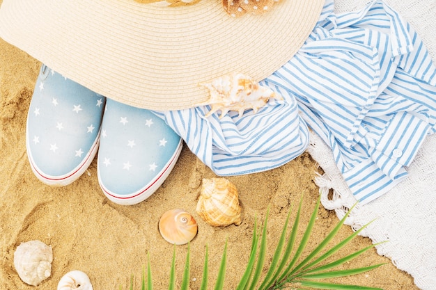 Bovenaanzicht van strandaccessoires op het zand. hoed, gestreept overhemd, schelpen, palmtakken en slip-on sneakers voor een tropische trip. zomervakantie.