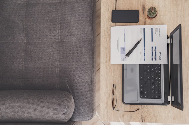 Bovenaanzicht van thuiskantoor werkplek. Laptop, factuur en bril op houten tafel, sofa