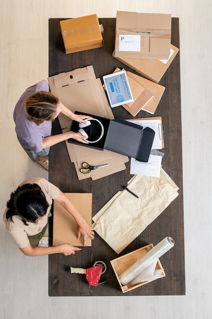 Bovenaanzicht van twee jonge vrouwelijke managers van een online winkel die aan tafel staan en bestellingen van klanten in dozen inpakken tijdens het werk in de opslagruimte store