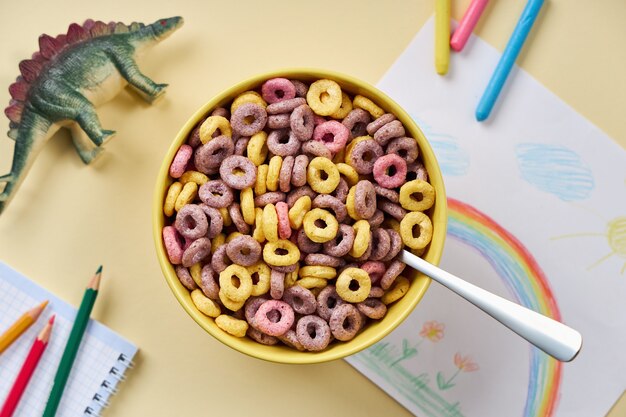 Bovenaanzicht van veelkleurige cornflakes in een gele kom met kinderspullen op een lichtgele achtergrond