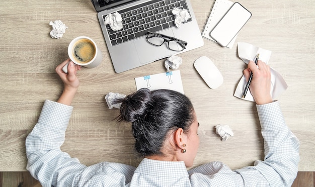 Bovenaanzicht van vermoeide, overwoekerde zakenvrouw, kantoormedewerker, liggend op het bureau met laptop, koffie en papieren.