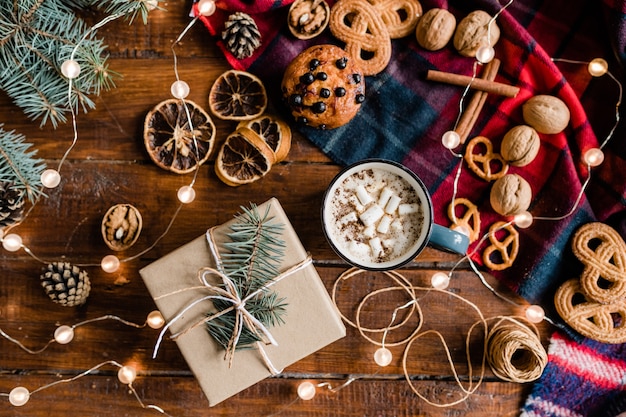 Bovenaanzicht van verpakt geschenk omgeven door warme drank, dennenappels, koekjes, walnoten, draden, naaldboom en slingers op houten tafel