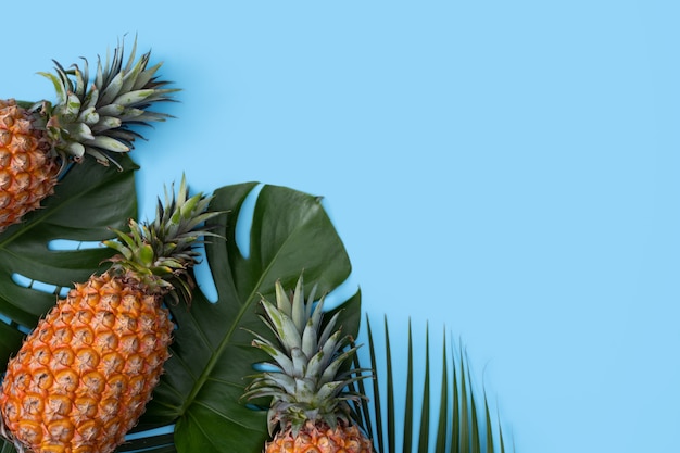 Bovenaanzicht van verse ananas met tropische palm en monsterabladeren op blauwe tafelachtergrond.