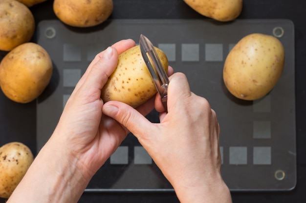 Bovenaanzicht van vrouw hand schillen van rauwe aardappel met dunschiller mes op de zwarte achtergrond