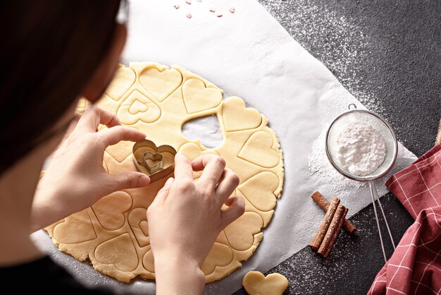 Bovenaanzicht van vrouw harten uit koekjesdeeg op keukentafel met poedersuiker en kaneel snijden