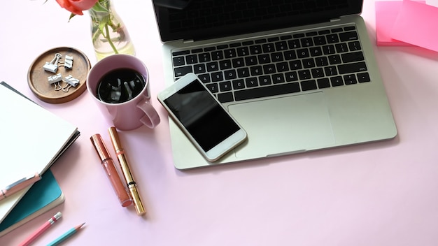 Bovenaanzicht van vrouw tafel met accessoires en vrouw cosmetische zetten. Computer laptop, smartphone, post-it, koffiekopje, cosmetica, notebook, clip en potplant Flat lag samen op roze tafel.