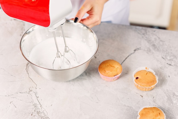 Bovenaanzicht van vrouwelijke banketbakker mengt een witte crème in een diepe metalen plaat met rode keukenmixer voor vers gebakken cupcakes.