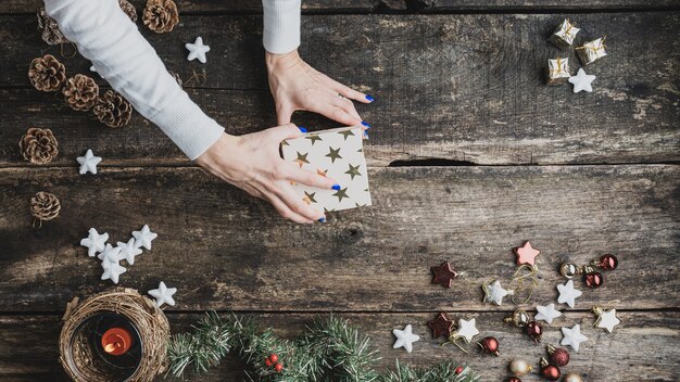 Bovenaanzicht van vrouwelijke hand die een vakantiegeschenkdoos sluit met gouden sterren in het midden van een rustiek houten bureau versierd met kerstversieringen en decoratie.