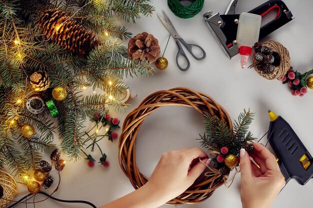 Bovenaanzicht van vrouwelijke handen maken een kerstkrans. Verpakte geschenken en rollen, sparren takken en gereedschappen op witte tafel. Voorbereiden op kerst- of nieuwjaarsvakantie