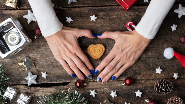 Bovenaanzicht van vrouwelijke handen met blauwe manicure die een hartvorm maakt rond een vakantiekoekje dat midden in de feestelijke kerstsfeer is geplaatst.