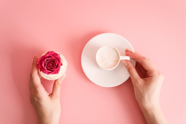 Bovenaanzicht van vrouwelijke handen met kopje koffie en cupcake versierd met roze knop roze achtergrond