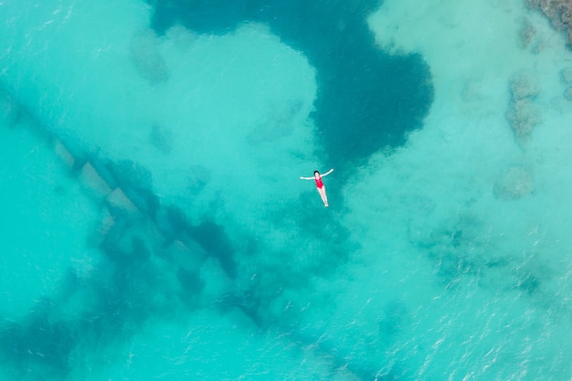 Bovenaanzicht vanuit de lucht van een vrouw die een rood zwempak draagt liggend op een transparant turquoise wateroppervlak op