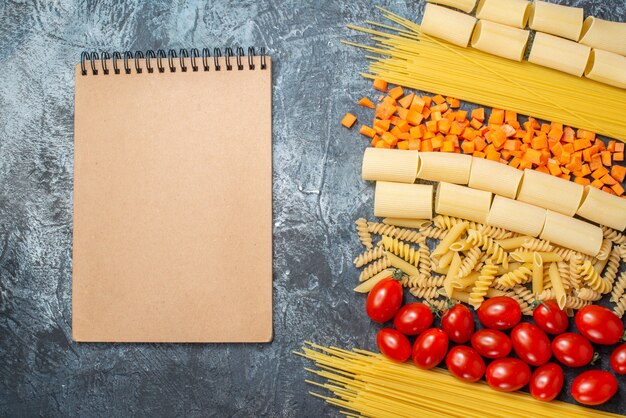 bovenaanzicht verschillende pasta's rigatoni spaghetti penne spiralen cherrytomaatjes gehakte wortel notitieblok op grijze achtergrond