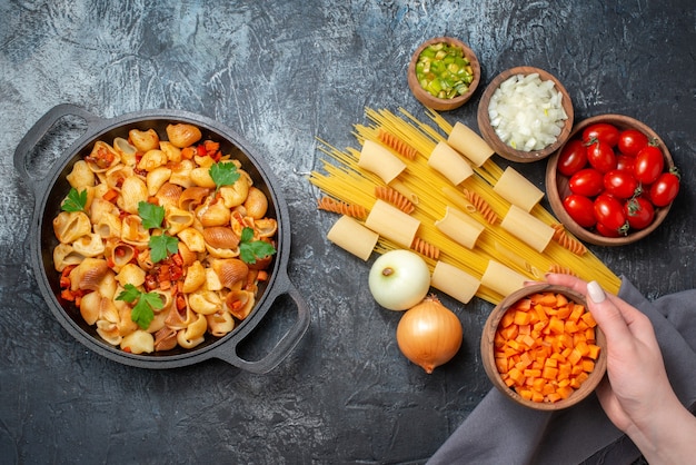 bovenaanzicht verschillende pasta's rigatoni spaghetti spiralen gehakte groenten in kommen smakelijke pasta in koekenpan kom met hakken wortel in vrouwelijke hand op grijze achtergrond